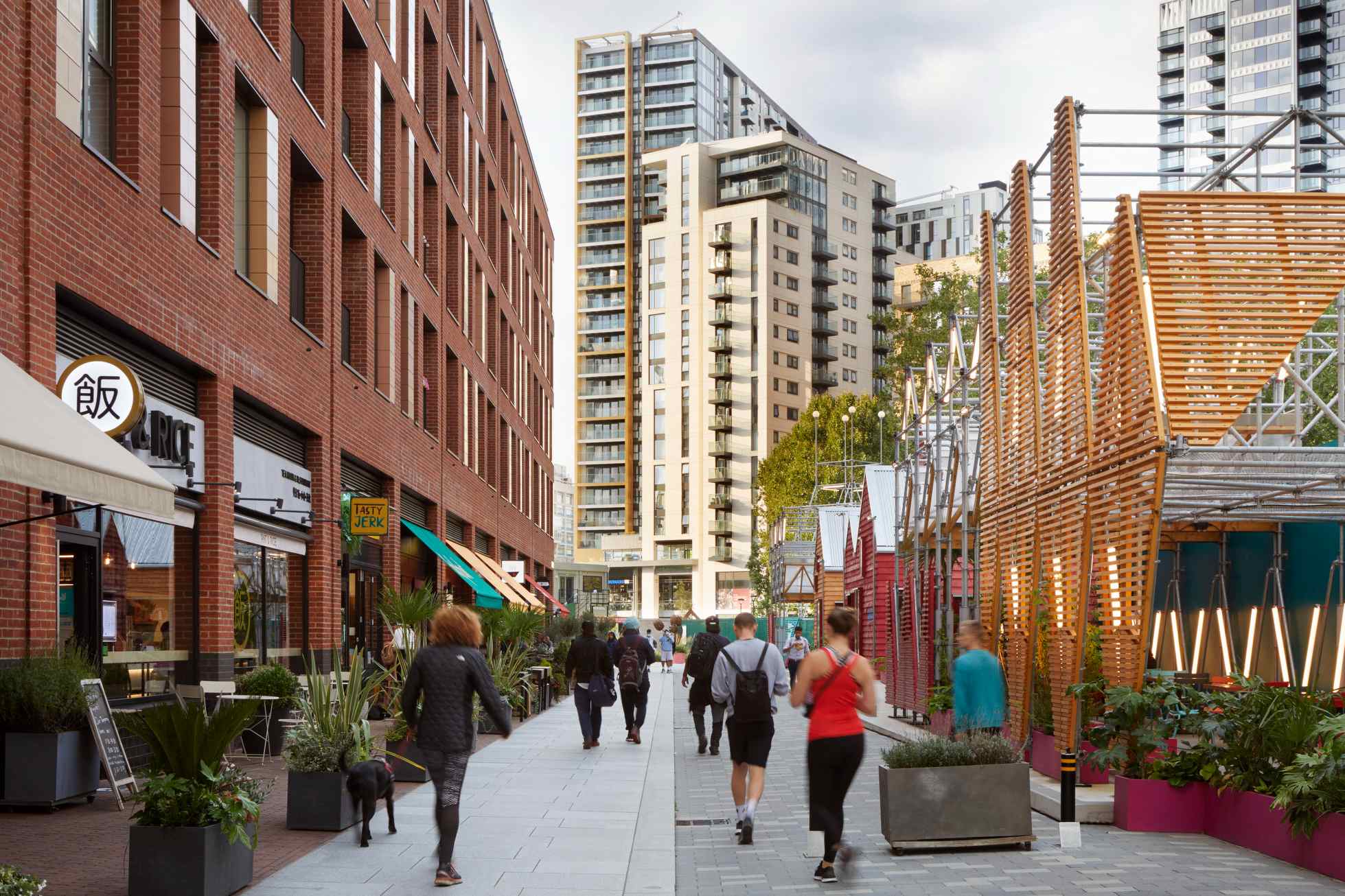 An image of Sayer Street at Elephant Park, with people walking down the street an red brick buildings on the left with the wooden structure of the Living Room on the right.