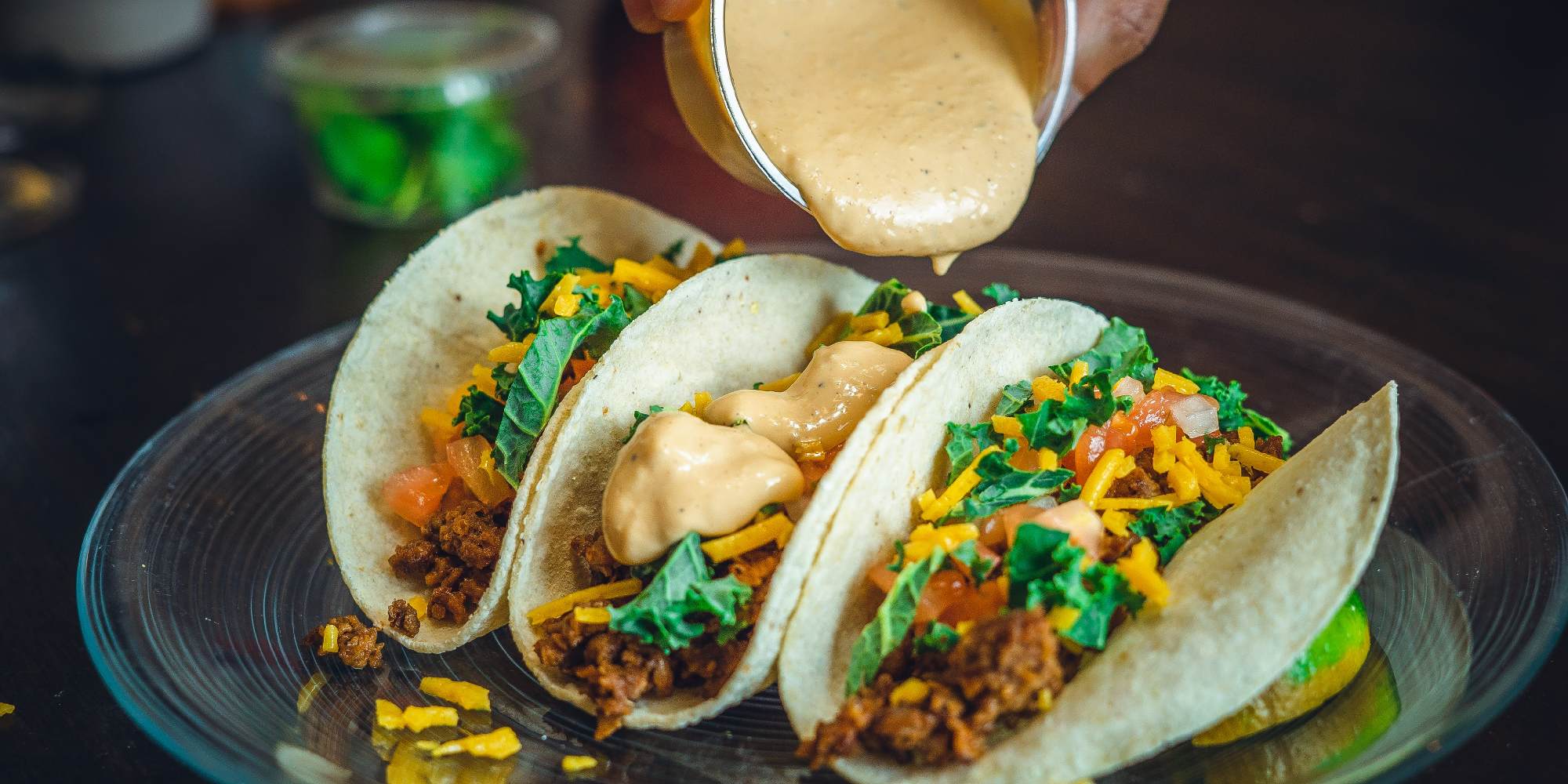 Three tacos on a plate with a hand pouring a sauce on top.