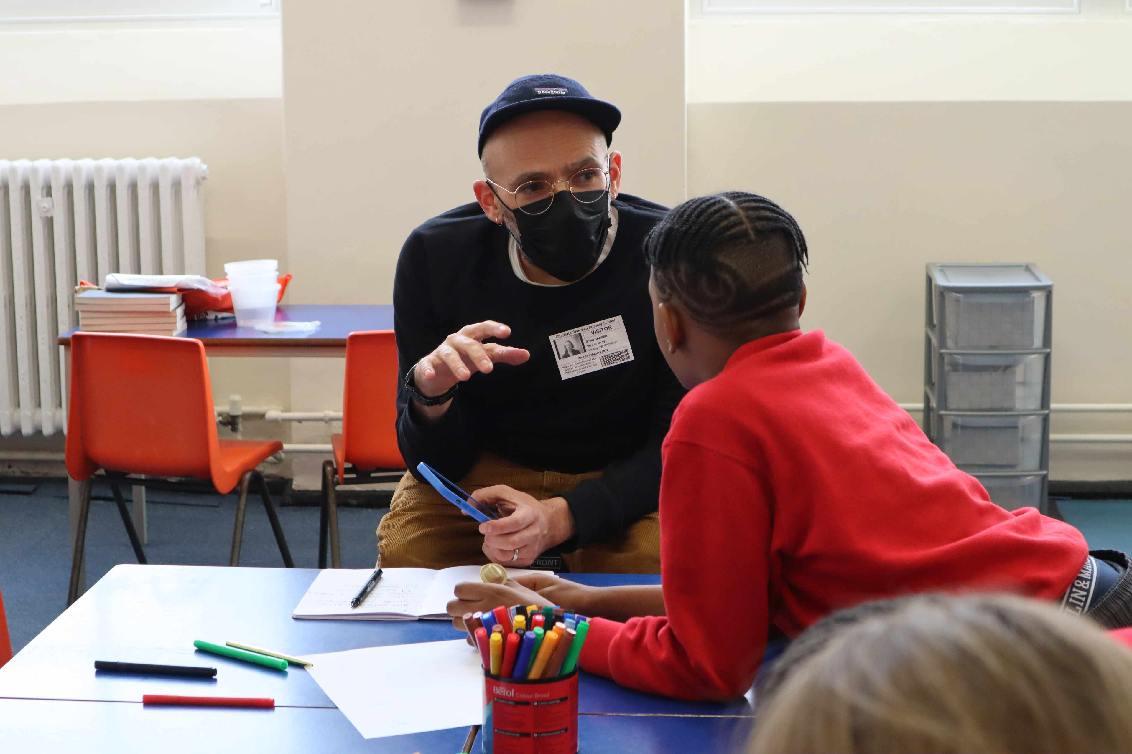 A primary school child wearing a red sweatshirt facing away from the camera and leaning across a table, talking to artist Ryan Gander who is wearing a black cap, glasses and a face mask.