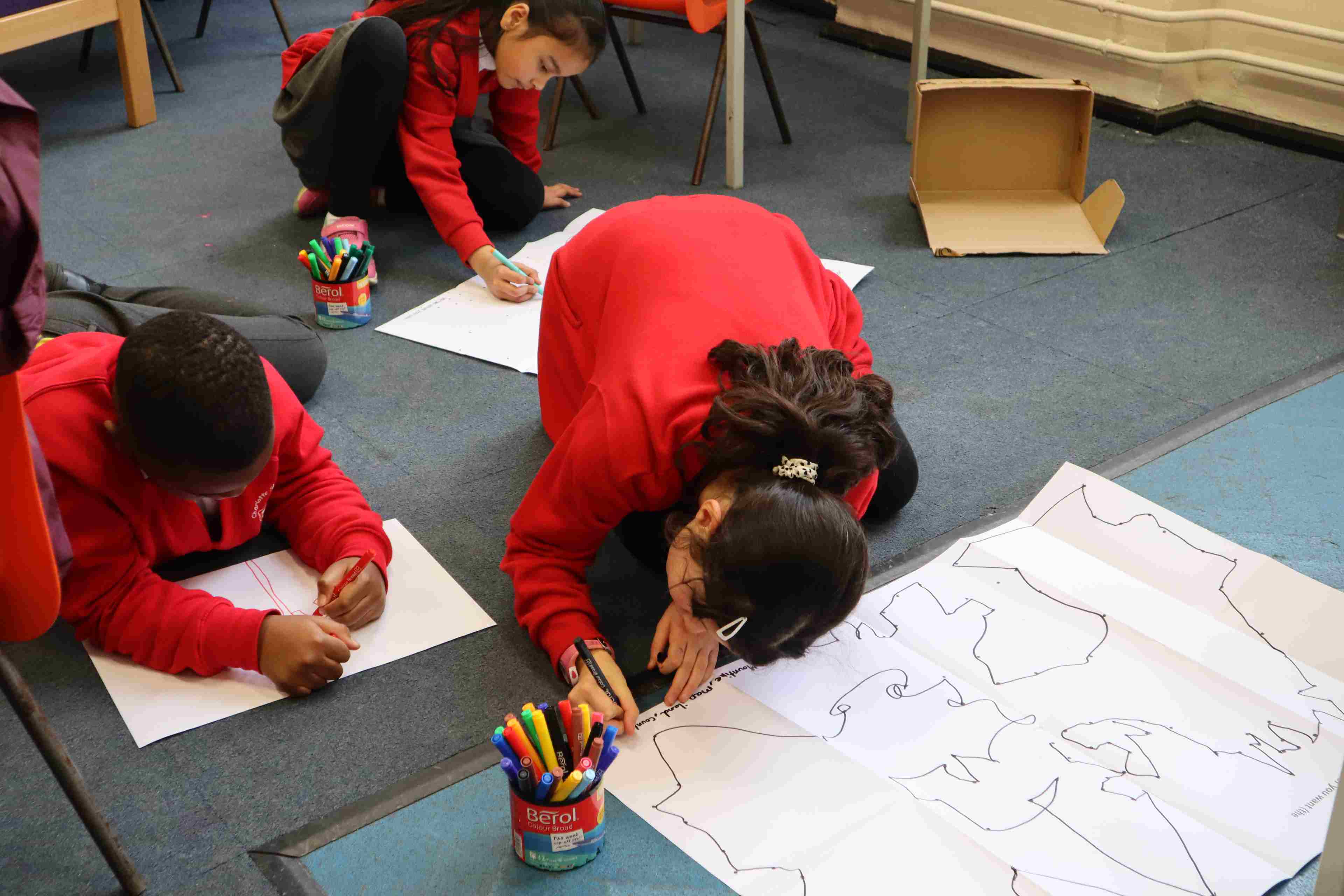 Primary school children wearing red sweatshirts  sitting on the floor colouring in pieces of paper.