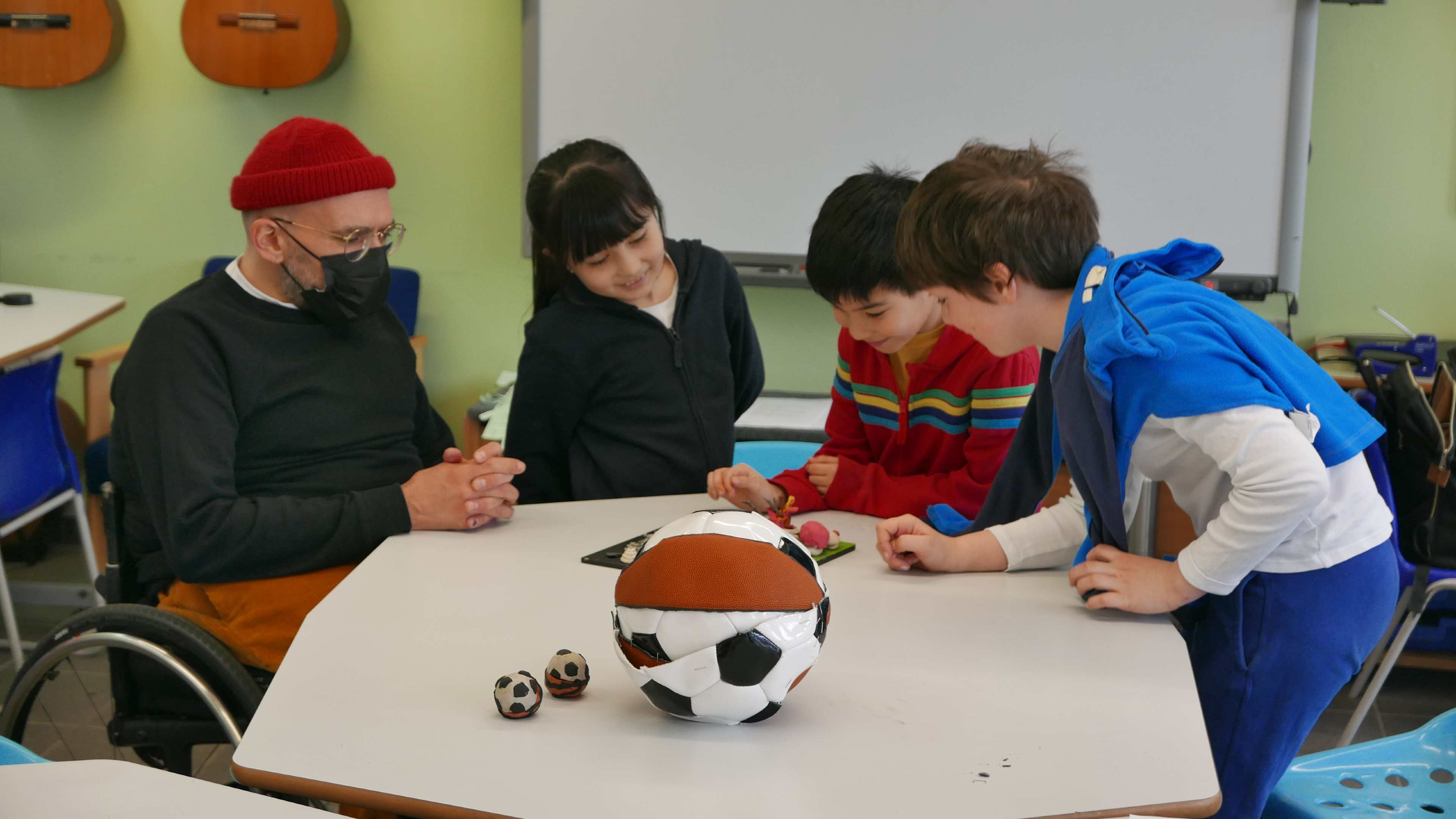 Three young children sitting around a table with artist Ryan Gander, with a football/basketball art piece in the centre.