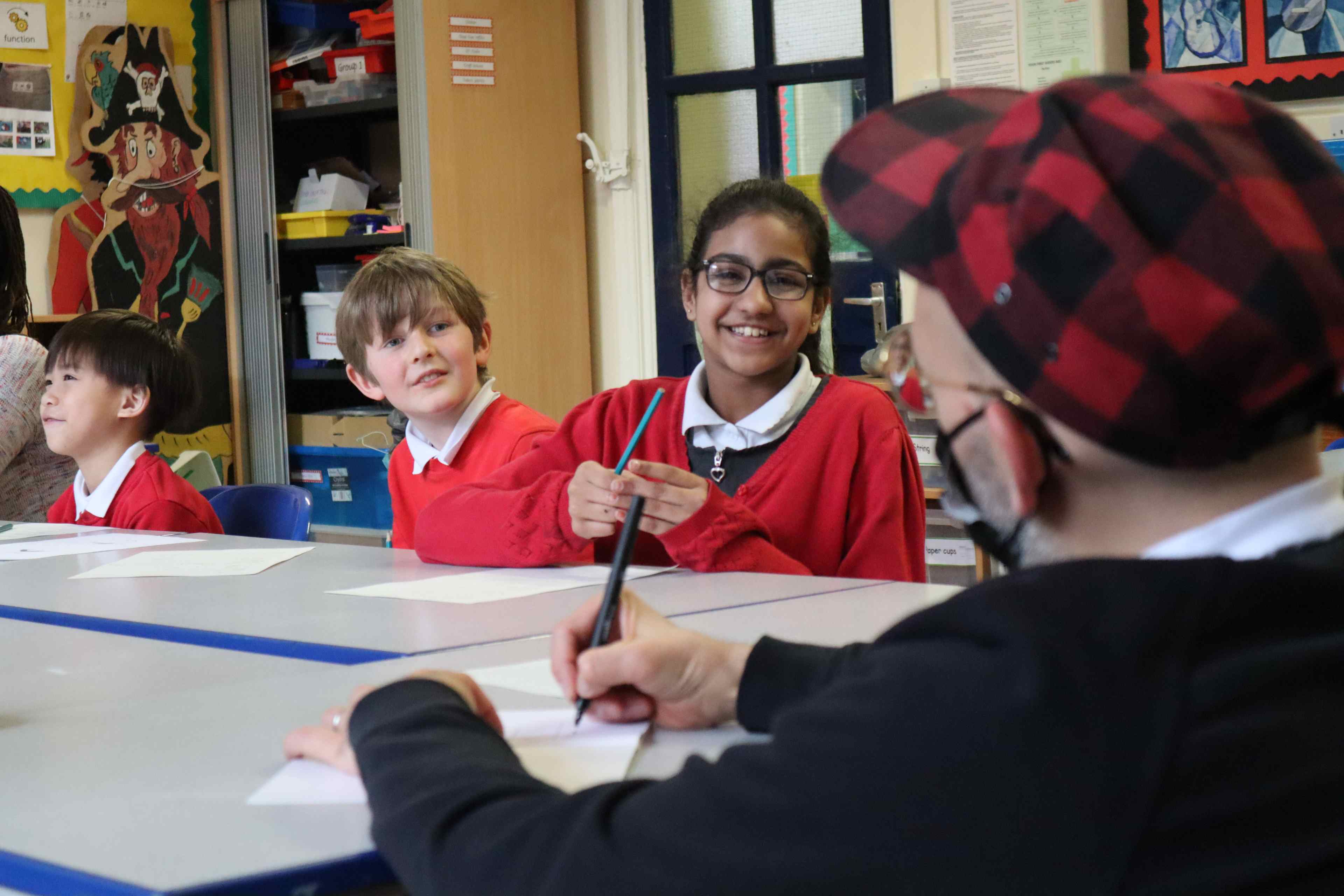 Three children wearing a red school uniform laughing with Ryan Gander at a table. Ryan Gander is facing away from the camera and is wearing a red and black tartan cap.